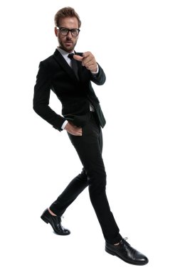 Siyah takım elbiseli kaşlarını çatmış işadamı elini cebine atıyor ve beyaz arka planda gururla yürüyor. 