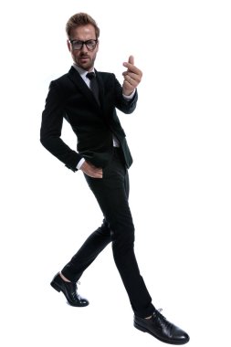 Siyah takım elbiseli kendine güvenen iş adamı elini cebine atıyor, parmaklarını ovuyor ve para jesti yapıyor, kaşlarını çatıyor ve beyaz arka planda şık bir şekilde yürüyor.