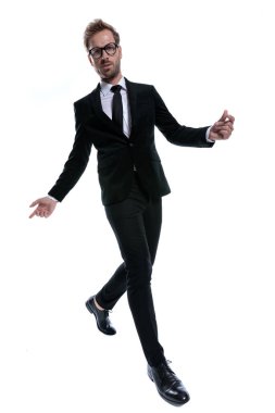 Siyah takım elbiseli, gözlüklü, kolları açık, beyaz arka planda tek başına yürüyen havalı bir adam.
