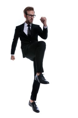 Siyah takım elbiseli somurtkan genç adam yan bakıyor, yumruklarını kaldırıyor ve beyaz arka planda tek başına yürüyor, tüm vücuduyla.