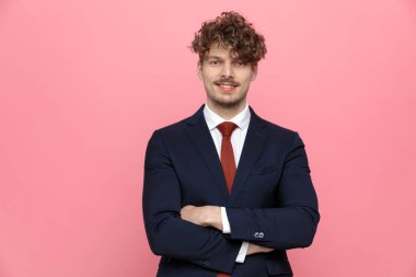 Lacivert takım elbiseli genç ve zarif bir iş adamı kollarını kavuşturup gülümsüyor ve pembe arka planda duruyor.