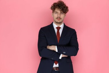 Kollarını kavuşturup pembe arka planda duran lacivert takım elbiseli ciddi bir genç adam.