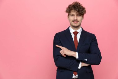 Güler yüzlü, lacivert takım elbiseli genç adam kollarını kavuşturup, parmaklarını sağa sola gösteriyor ve pembe arka planda gülümsüyor.