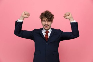 heyecanlı mavi takım elbiseli genç işadamı yumruklarını havaya kaldırmış, gülüyor ve zaferi kutluyor, pembe arka planda duruyor.