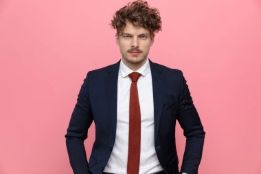 Lacivert takımlı zarif genç işadamı ceplerinde el ele tutuşup pembe arka planda poz veriyor.