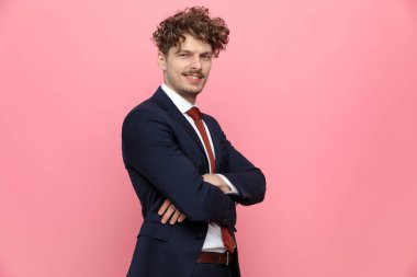 Mavi lacivert takımlı, kollarını kavuşturup gülümseyen, ayakta duran ve pembe arka planda poz veren mutlu zarif adamın yan görüntüsü.