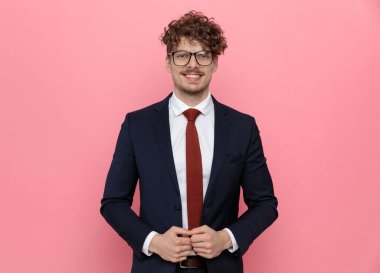 Lacivert takım elbiseli mutlu genç işadamı gözlük takıyor, parmaklarına dokunuyor, takım elbise ayarlıyor ve pembe arka planda gülümsüyor.