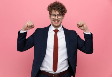 Deniz mavisi takımlı heyecanlı genç adam gözlük takıyor, yumruklarını havaya kaldırıyor ve zaferi kutluyor, pembe arka planda duruyor.