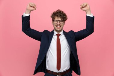 Lacivert takımlı gururlu iş adamı gözlüklü, yumruklarını havaya kaldırmış ve pembe arka planda zafere tezahürat yapıyor.