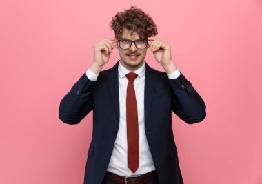 Lacivert takımlı mutlu genç işadamı gözlüklerini ayarlıyor ve pembe arka planda gülümsüyor.