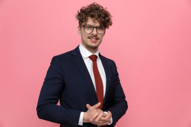 Lacivert takımlı genç işadamı gözlük takıyor, avuç içi ovuyor ve hazırlanıyor, pembe arka planda gülümsüyor.