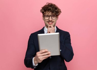 Lacivert takım elbiseli mutlu genç işadamı gözlük takıyor. El ele tutuşuyor ve düşünüyor. Pembe arka planda gülümsüyor.