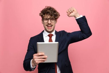 heyecan verici mavi takım elbiseli genç işadamı, gözlük takıyor, yumruklarını havaya kaldırıyor ve zaferi kutluyor, pembe arka planda bağırıyor ve gülümsüyor.