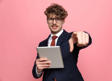 Lacivert takım elbiseli zarif genç işadamı. Gözlük takıyor. Hesap tutuyor ve pembe arka planı işaret ediyor.