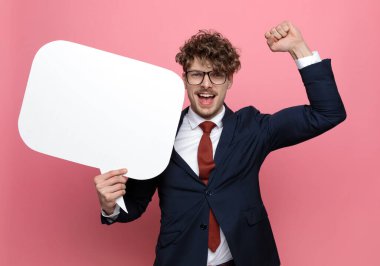 Lacivert takımlı mutlu genç işadamı gözlük takıyor, konuşma baloncuğu tutuyor, yumruklarını havaya kaldırıyor ve zaferi kutluyor, pembe arka planda gülüyor ve çığlık atıyor.