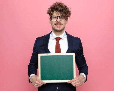 Lacivert takım elbiseli mutlu genç işadamı. Tahtayı sunuyor ve pembe arka planda gülümsüyor.