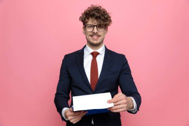 Lacivert takım elbiseli, elinde gözlük olan, pembe arka planda sunan ve gülümseyen genç bir işadamı.