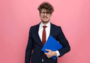 Lacivert takım elbiseli, gözlüklü, elinde not panosu olan ve pembe arka planda gülümseyen mutlu bir iş adamı.