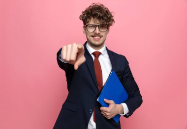Lacivert takım elbiseli, elinde pano olan ve pembe arka planı işaret eden mutlu genç bir adam.
