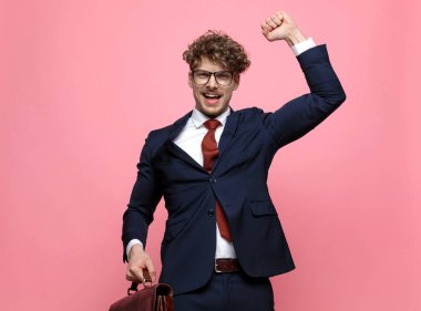 Lacivert takımlı mutlu bir adam bavul tutuyor, yumruklarını havaya kaldırıyor ve pembe arka planda zaferi kutluyor.