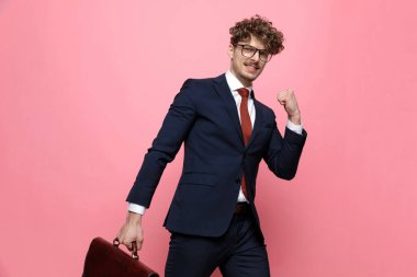 Lacivert takımlı genç adam bavulu tutuyor, yumruklarını havaya kaldırıyor ve zaferi kutluyor, pembe arka planda yürüyor.