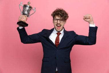 Deniz mavisi takım elbiseli heyecanlı genç bir manken gözlük takıyor, kupa tutuyor, yumruklarını havaya kaldırıyor, çığlık atıyor ve pembe arka planda zaferi kutluyor.