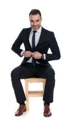 Gülen iş adamı beyaz stüdyo arka planında bir sandalyede otururken ceketinin düğmelerini açıyor.