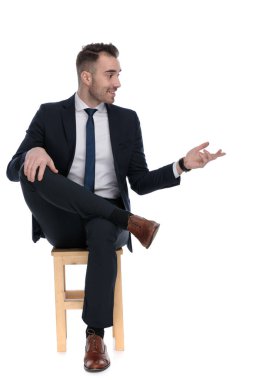 Mutlu iş adamı gülümsüyor ve konuşuyor. Beyaz stüdyo arka planında bir sandalyede otururken el kol hareketi yapıyor.