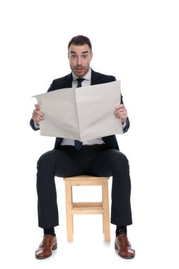 Beyaz stüdyo arka planında bir sandalyede otururken gazete okuyan şaşkın iş adamı.