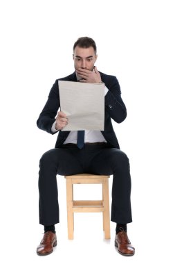 Gazete okuyan, nefes nefese kalan ve beyaz stüdyo arka planında bir sandalyede otururken ağzını kapatan şaşkın iş adamı.