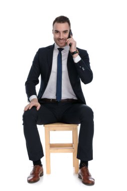 Mutlu iş adamı telefonda konuşuyor ve beyaz stüdyo arka planında bir sandalyede otururken gülümsüyor.