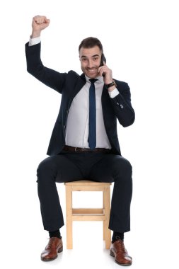 Neşeli iş adamı telefonda konuşuyor ve kutlama yapıyor. Beyaz stüdyo arka planında bir sandalyede otururken gülüyor.