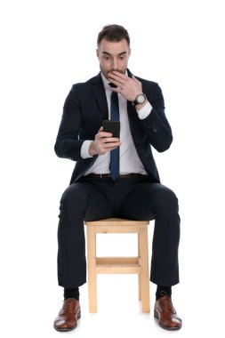 Şaşırmış iş adamı elinde telefon tutuyor ve nefes nefese, beyaz stüdyo arka planında bir sandalyede otururken ağzını kapatıyor.