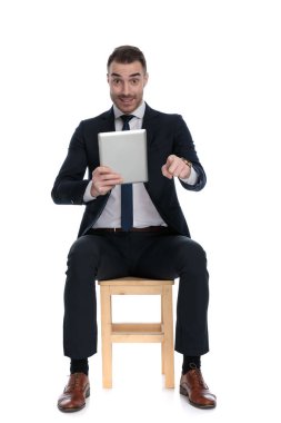 Pozitif iş adamı, beyaz stüdyo arka planında bir sandalyede otururken gülümseyerek ve tablet tutarak öne bakıyor.