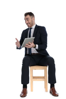 Heyecanlı iş adamı beyaz stüdyo arka planında bir sandalyede otururken tablet ve el kol hareketi yapıyor.