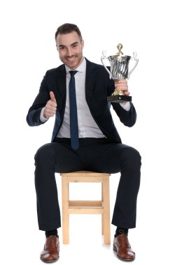 Pozitif iş adamı, beyaz stüdyo arka planında bir sandalyede otururken gülümsüyor ve kupa tutuyor.