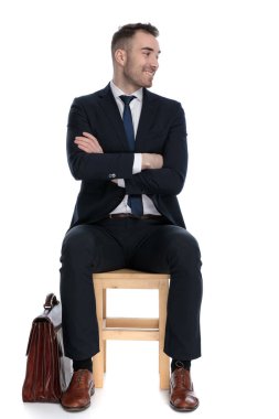 Pozitif iş adamı beyaz stüdyo arka planında bir sandalyede otururken evrak çantasının yanında el ele tutuşup gülüyor.