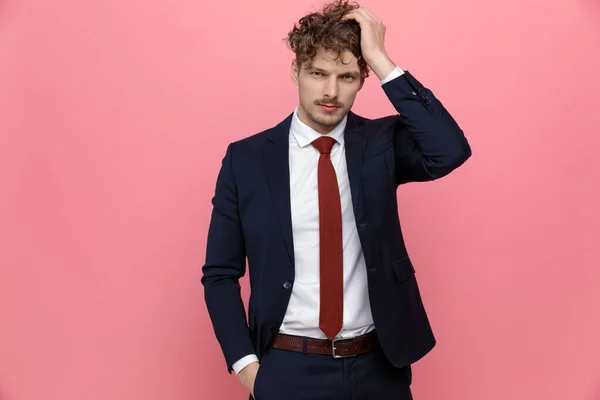 clueless young businessman in navy blue suit holding hand in pocket and scratching head, thinking and standing on pink background