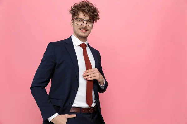 happy young businessman in navy blue suit wearing glasses, holding hands in pockets and fixing tie, smiling on pink background