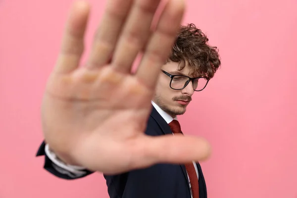 upset young businessman in navy blue suit, looking down and making stop gesture on pink background