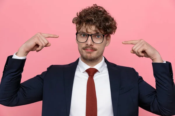 young elegant fashion man in navy blue suit wearing glasses and pointing fingers to head, standing on pink background