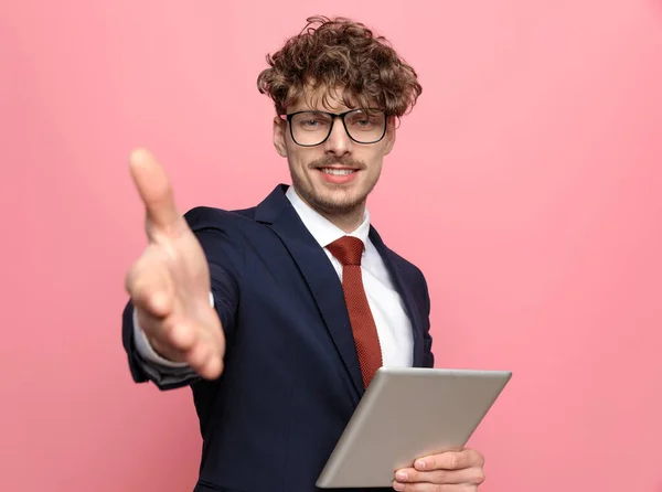 elegant young fashion man in navy blue suit wearing glasses holding tab and shaking hand, welcoming and smiling on pink background