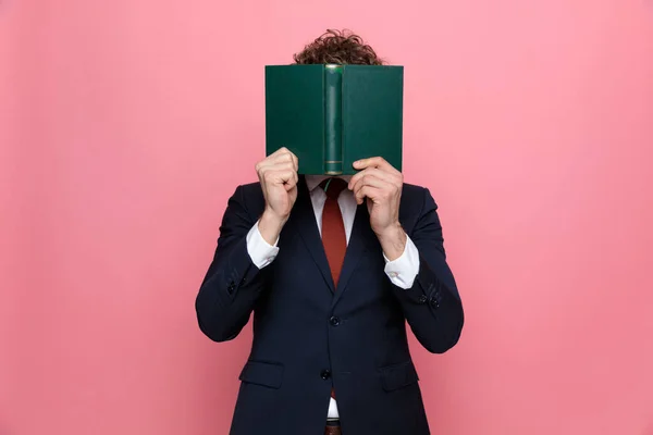 elegant businessman in navy blue suit hiding behind book on pink background