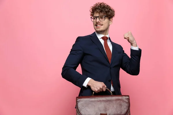 ネイビーブルーのスーツに身を包んだ実業家がスーツケースを持ち横を向いてピンクを背景に拳を握り勝利を祝う — ストック写真