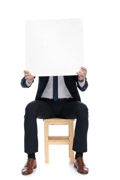 年轻的商人坐在白色画室背景的椅子上 用空白的广告牌盖住脸 — 图库照片