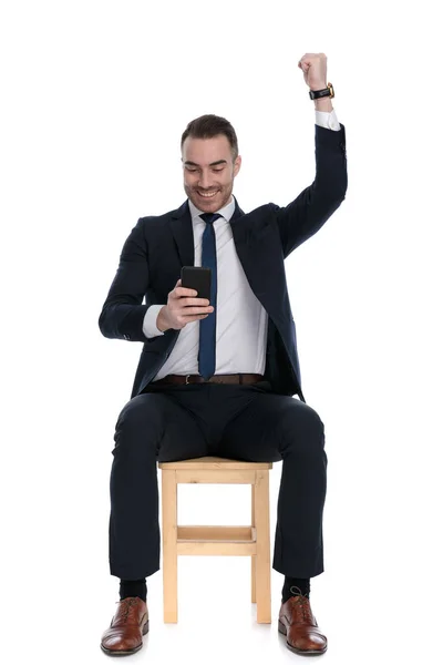 兴高采烈的商人在电话里发短信 手握拳头 坐在白色画室的椅子上庆祝 — 图库照片