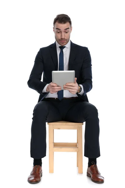 感兴趣的商人坐在白色工作室背景的椅子上 一边看着平板电脑 一边微笑 — 图库照片