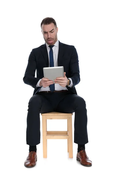 持平板电脑的持不同政见的商人 坐在白色工作室背景的椅子上 露出舌头皱着眉头 — 图库照片