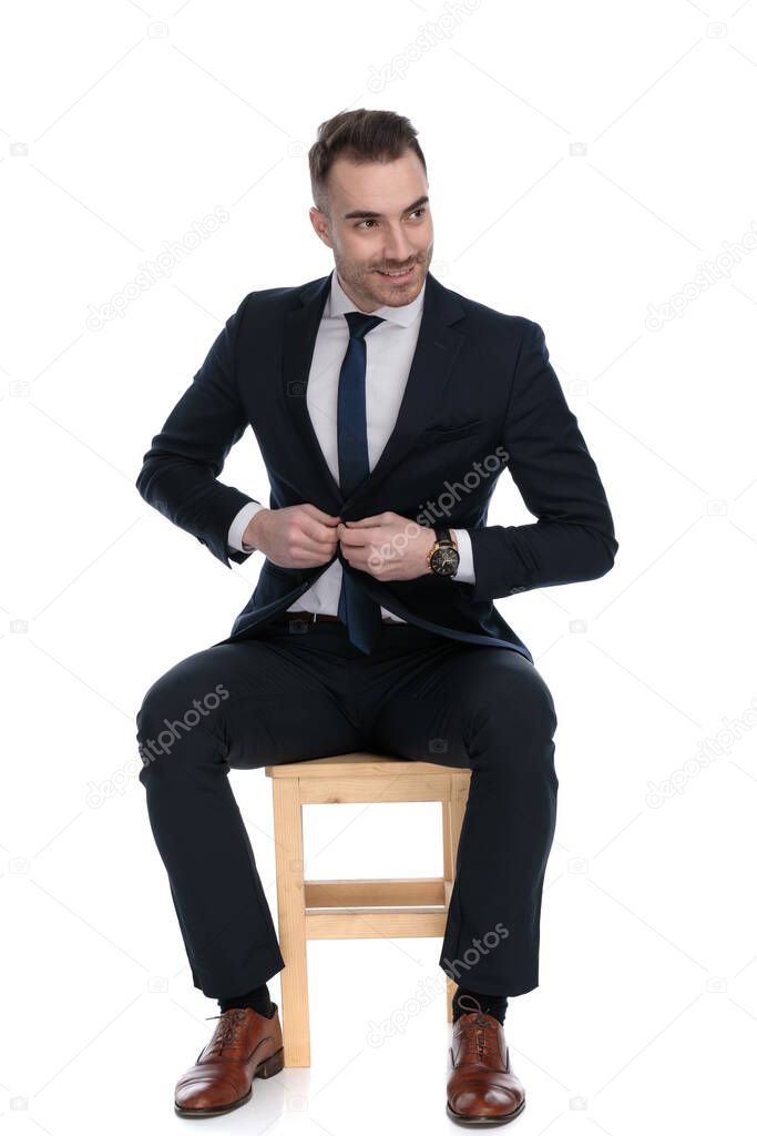 Happy businessman unbuttoning his jacket and looking away while sitting on a chair on white studio background
