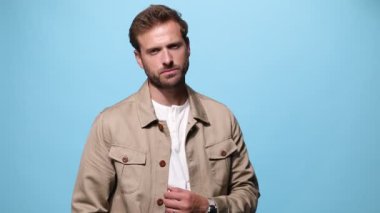 Çekici, sıradan bir adam, yüzünde bir gülümsemeyle ceketini düzeltiyor, kollarını göğsünde kavuşturuyor, yana dönüyor ve mavi arka plana bakıyor.